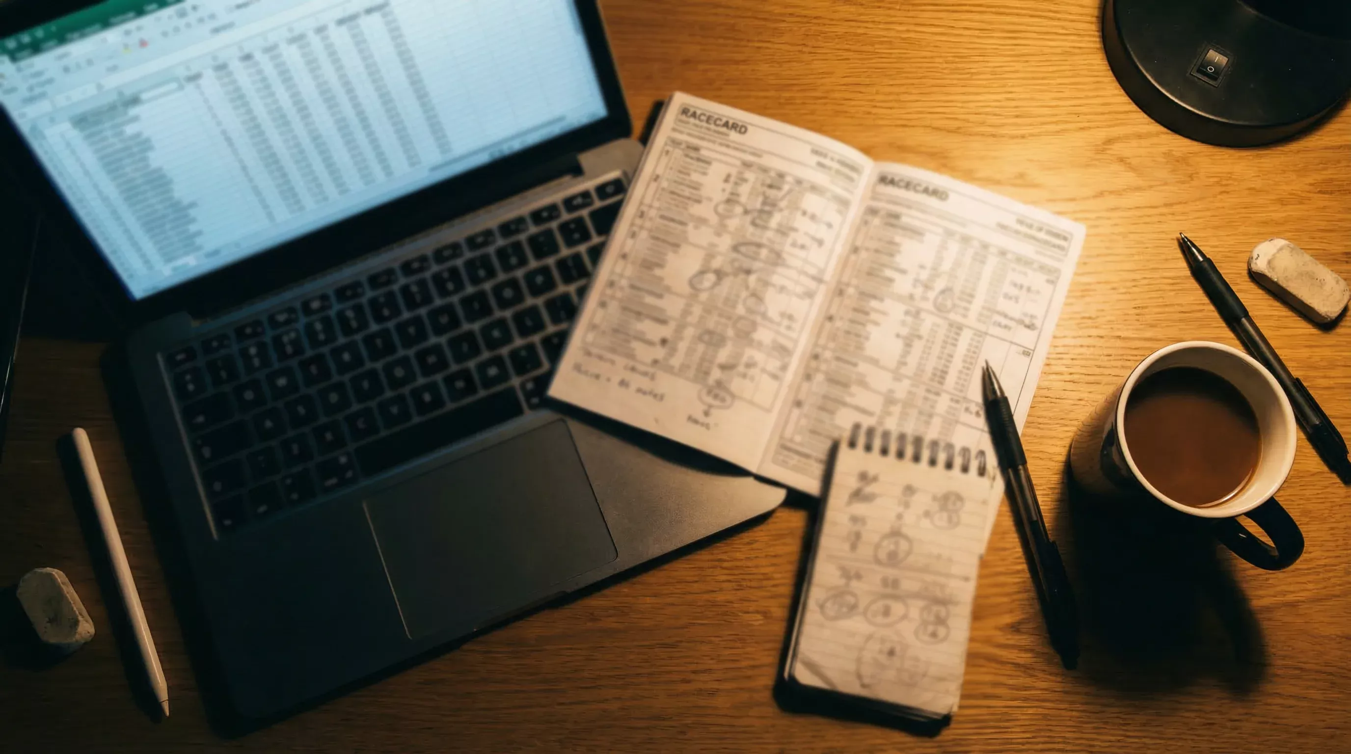 Person reviewing greyhound form data on a laptop screen with a racecard and notepad beside it