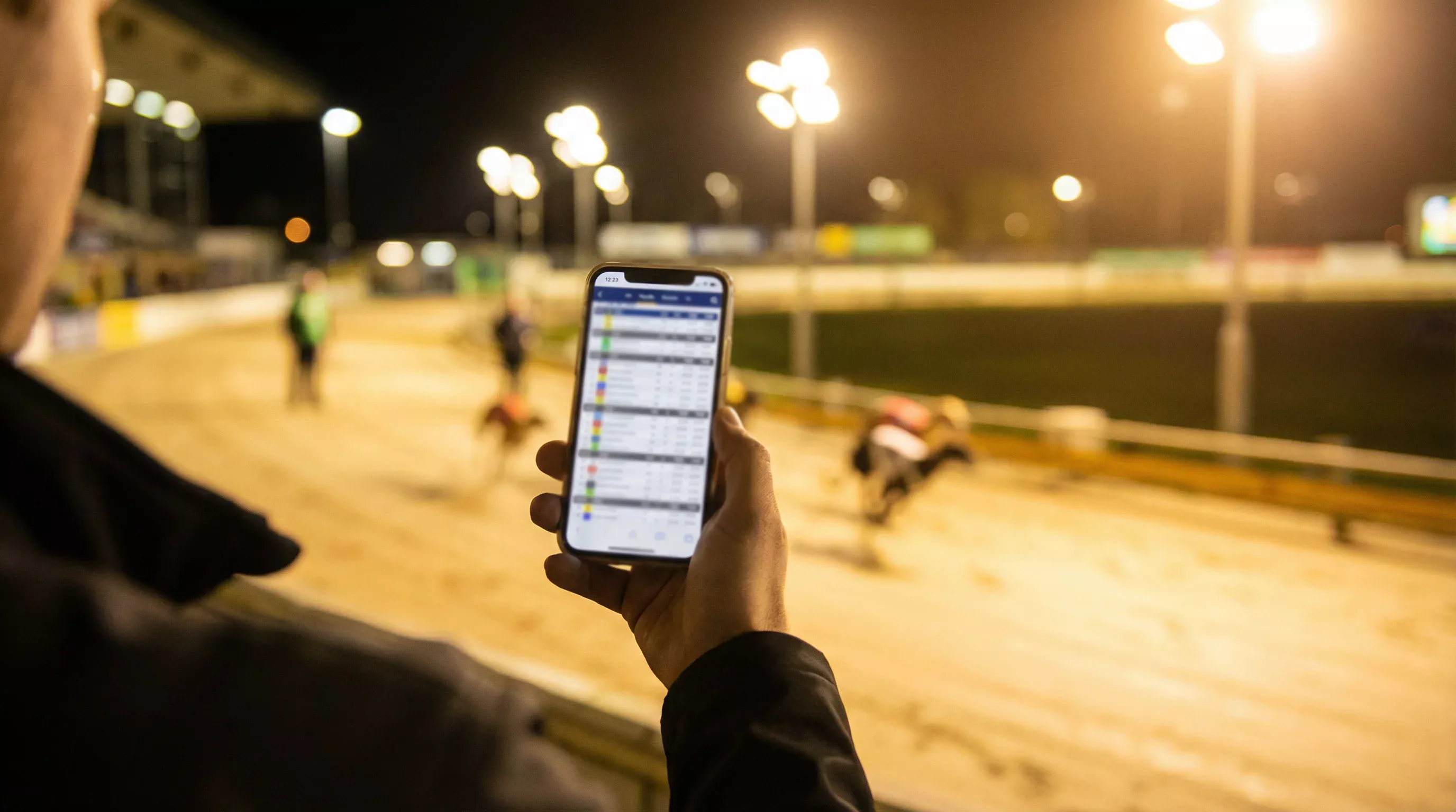 Smartphone screen displaying live greyhound race results with a sand track blurred in the background